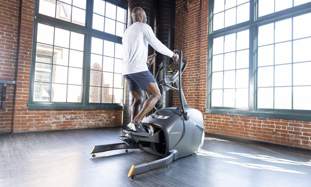 Man training on an elliptical machine in a bright, sunlit room with large windows.